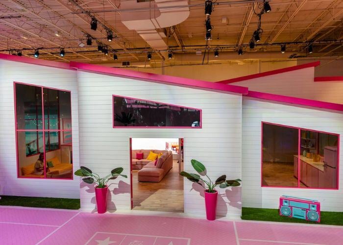  A pink and white Barbie Dreamhouse with a large living room visible through the open front door. There are potted plants on either side of the door and a boombox to the right.