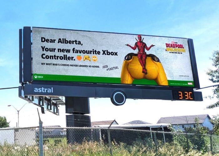 A large billboard featuring Deadpool, the superhero, sitting on a giant Xbox controller. The text on the billboard reads "Dear Alberta, Your new favorite Xbox Controller. MY BEST BUB'S CHEEKS NEVER LOOKED SO GOOD. XOXO DEADPOOL" and "XBOX." The billboard is also advertising the upcoming Deadpool and Wolverine film. This is a film marketing campaign for the movie.