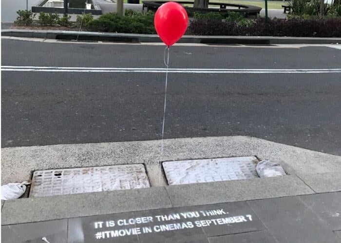 A red balloon tied to a string is floating above a storm drain on a city street. Chalk graffiti on the sidewalk reads "IT IS CLOSER THAN YOU THINK" and "#ITMOVIE INCINEMAS SEPTEMBER 7." This is a guerrilla marketing campaign for the film "It" (2017).