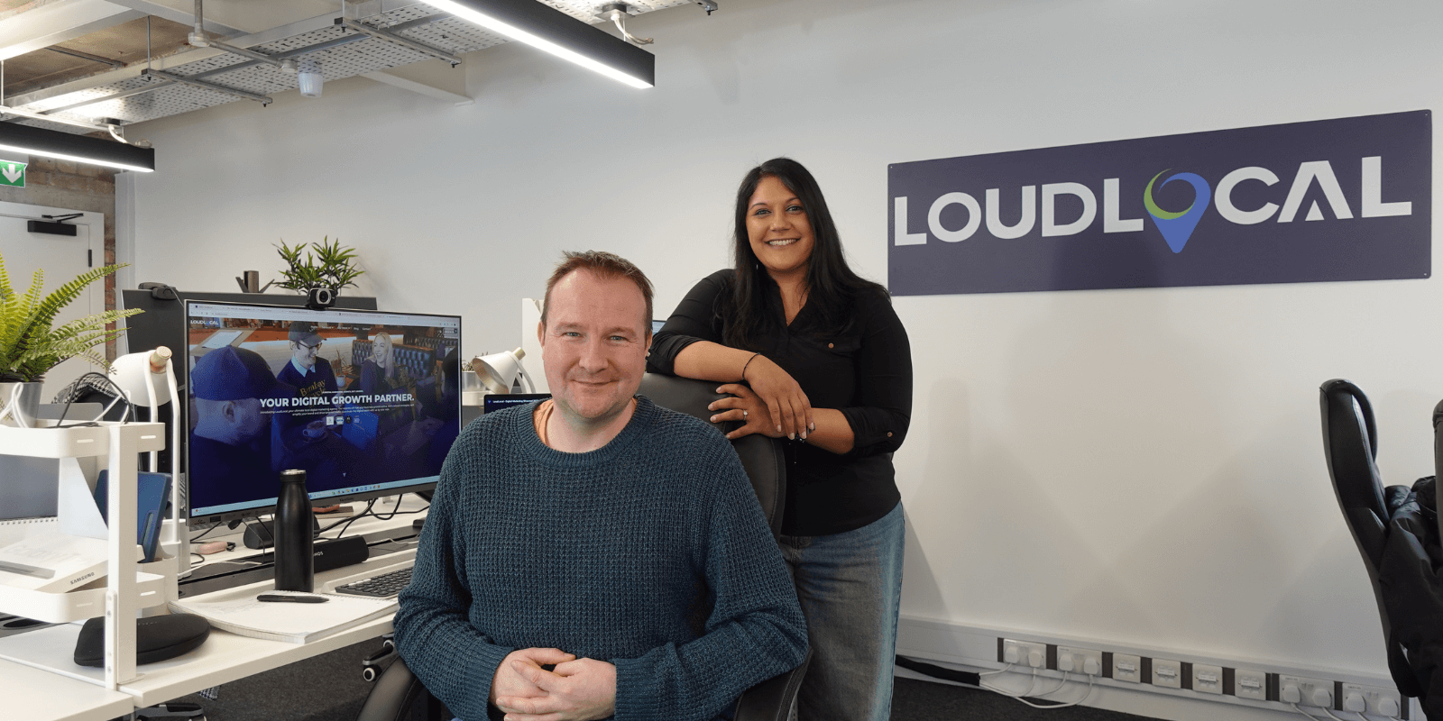 A picture of our 2 founders, Leon Hidderley and Priya Pandit in the new office, with the LoudLocal sign behind and a computer open on the LoudLocal website