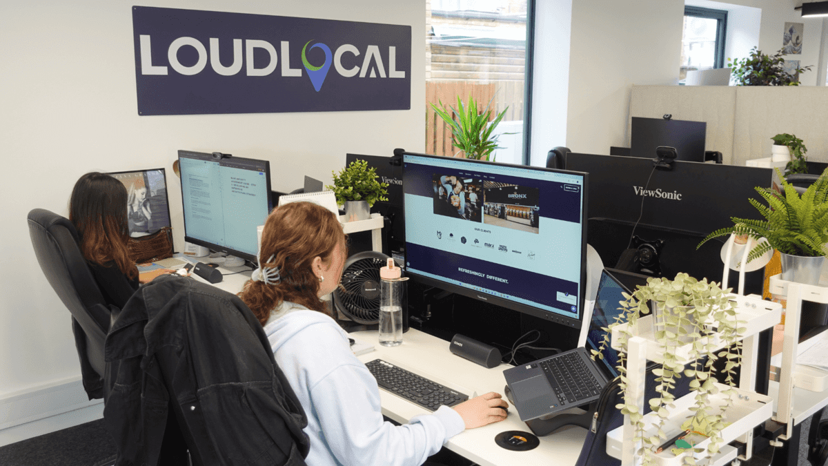 The LoudLocal team.  Shown in the picture is Mia and Tanya, two marketing assistants sat at their desks looking at their screens.  In the background their is a dark blue loudlocal logo.  The desks are white and Mia and Tanya at sat on black chairs.  Tanya has short dark straight hair and mia has curly brown hair wrapped in a bobble.  The office has lots of plants too. 